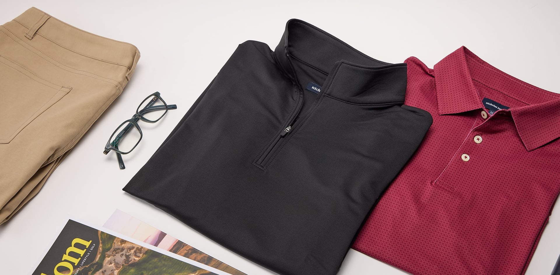 Black half-zip pullover, red polo shirt, beige pants, and eyeglasses on a white surface.